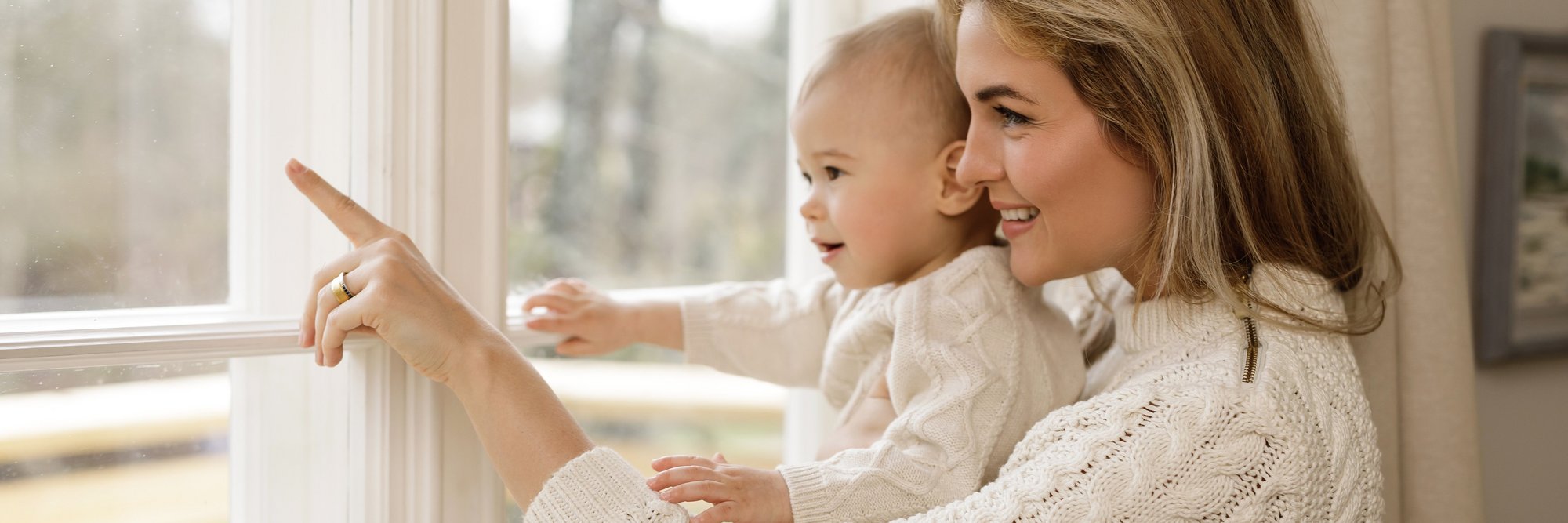 Mutter zeigt mit Baby auf etwas vor dem Fenster. Das Bild zeigt eine Mutter, die mit ihrem Baby vor einem Fenster steht und auf etwas zeigt. Beide wirken neugierig und entspannt.