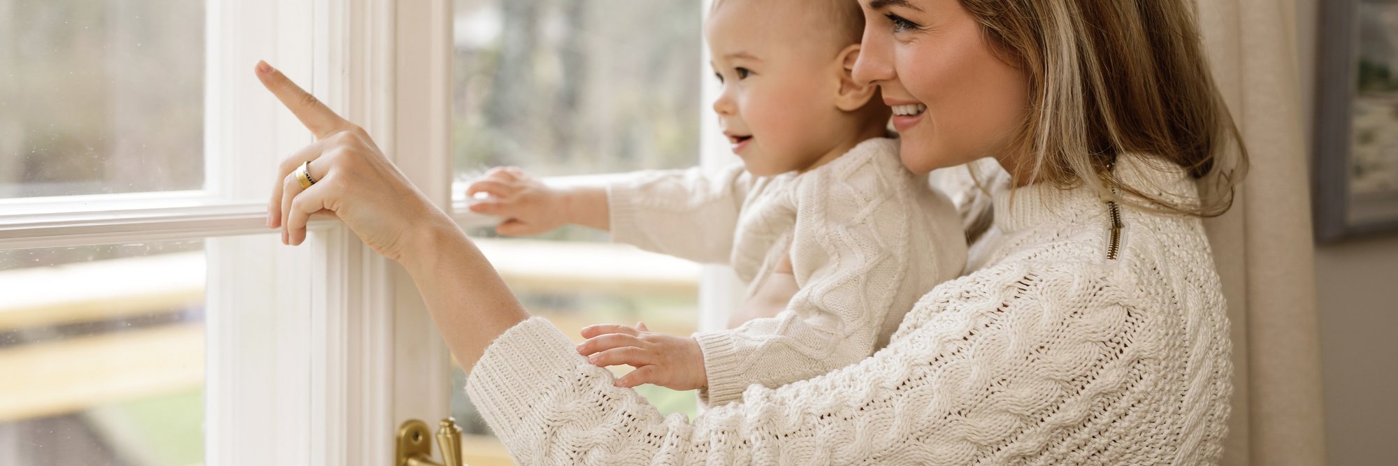 Das Bild zeigt eine Mutter, die mit ihrem Baby vor einem Fenster steht und auf etwas zeigt. Beide wirken neugierig und entspannt.