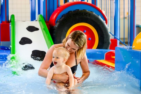  Eine Frau und ein Kind spielen fröhlich im Wasserpark, umgeben von spritzendem Wasser und bunten Rutschen.