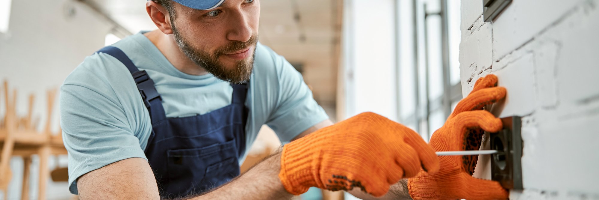Handwerker installiert Steckdose an der Wand. Das Bild zeigt einen Handwerker, der in Arbeitskleidung und mit Handschuhen an einer Wand eine Steckdose installiert.