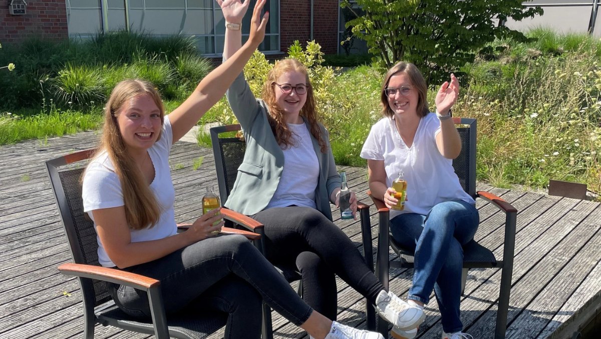 Das Bild zeigt drei Personen, die sich draußen auf einer Terrasse befinden und freundlich winken.