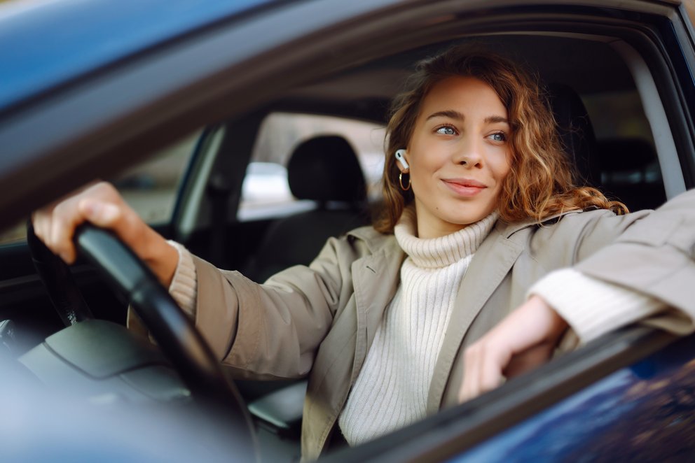 Eine Frau im Auto hinter dem Lenkrad sitzend, sie hat einen Arm aus dem Fenster gelehnt und schaut aus dem Fenster.