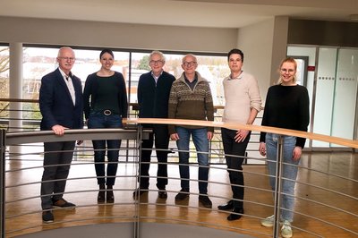 sechs Personen, die gemeinsam in einem modernen Innenraum an einem Geländer stehen und in die Kamera lächeln