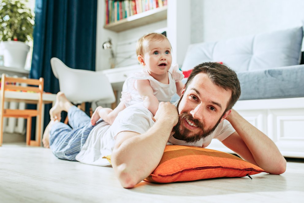Entspannter Vater mit spielendem Kind auf dem Rücken. Das Bild zeigt einen Vater, der auf dem Boden liegt und sich entspannt. Auf seinem Rücken sitzt sein kleines Kind, das fröhlich spielt.