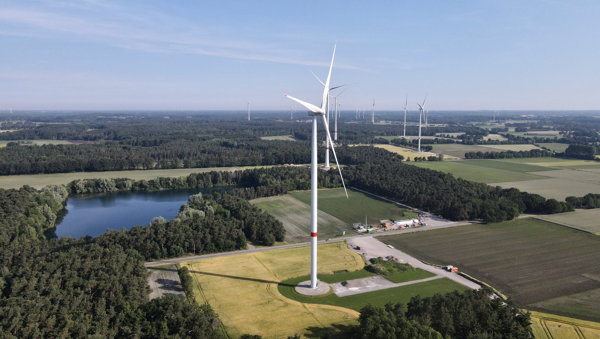 Luftaufnahme von Windkraftanlagen auf einem weiten, grünen Feld, umgeben von Bäumen und einem See.