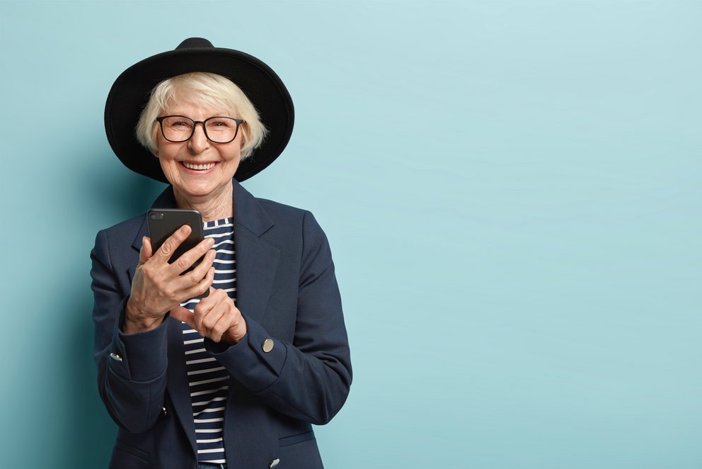 Eine ältere Frau blickt lachend in die Kamera, sie trägt einen Hut und hält ein Handy in der Hand. Pastellblauer Hintergrund.
